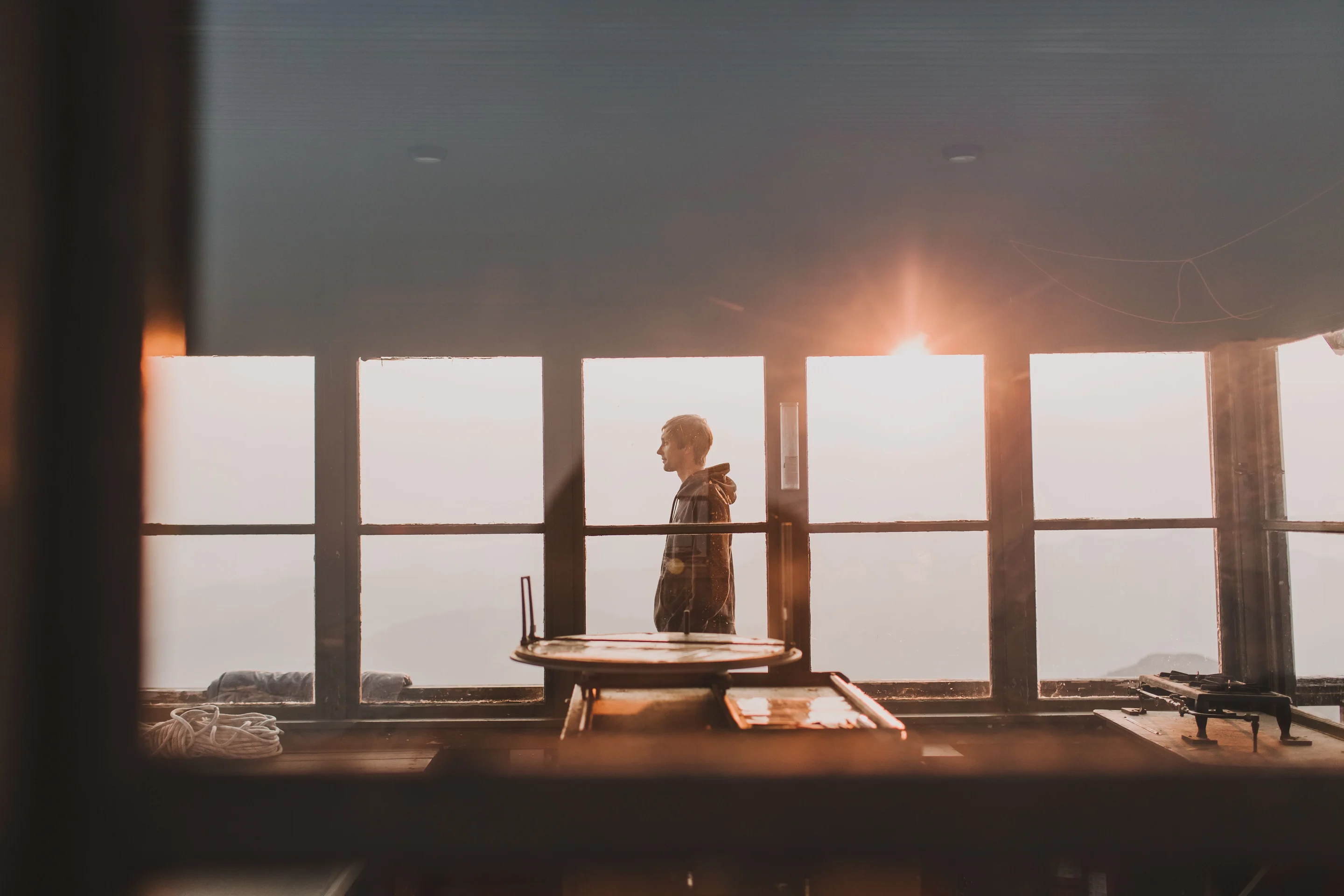 A man standing in a mountain fire-lookout at sunset, looking out over the hills — a quiet moment of freedom and peace.